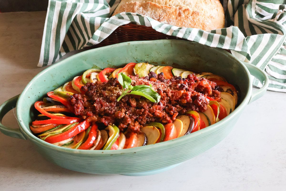 Ratatatouille with meat in a green casserole dish with crusty bread in a green striped towel.