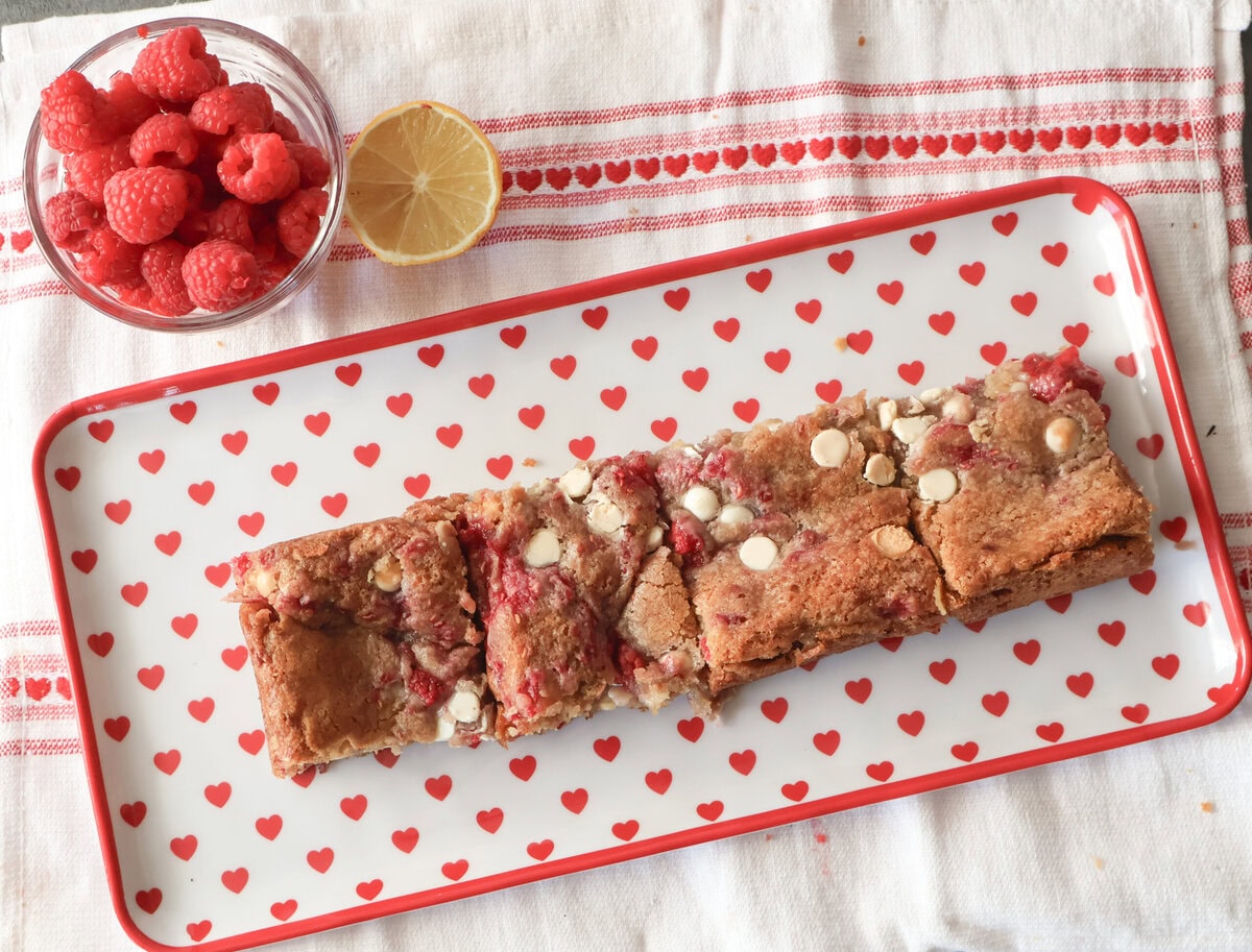 White chocolate raspberry blondies on a heart plate with raspberries and lemon.