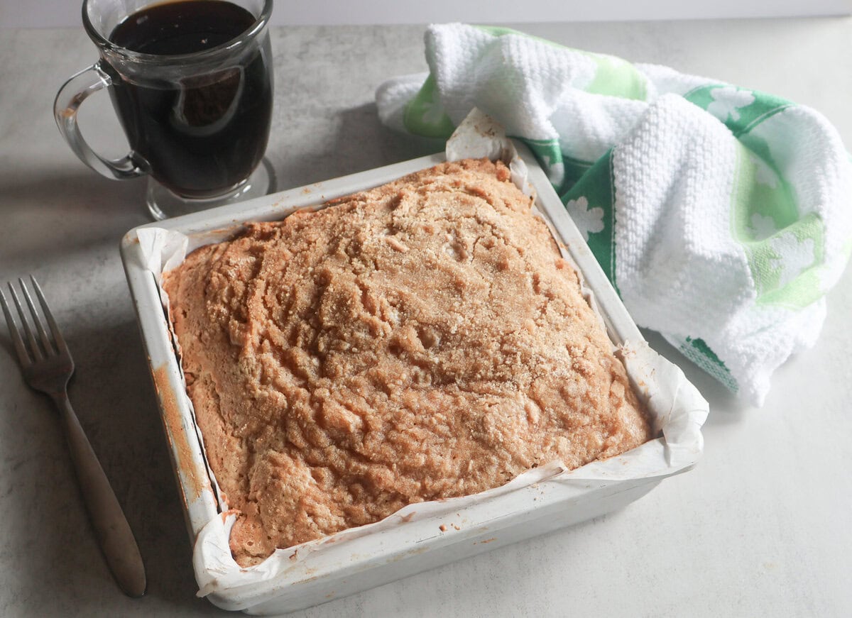 Baked Irish Cream Coffee Cake in a square pan with a cup of coffee and a green and white tea towel.