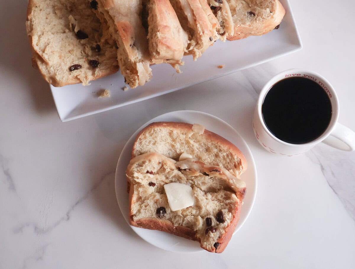 Slice of Spiced Hot Cross Buns Loaf on a white plate with a cup of coffee.