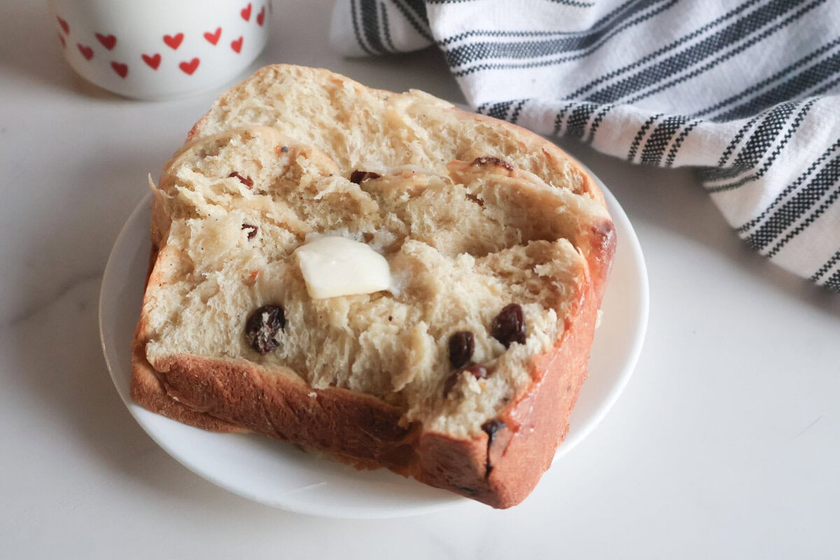 Slice of Hot Cross Buns Loaf on a white plate with a pat of butter and a gray and white striped tea towel.