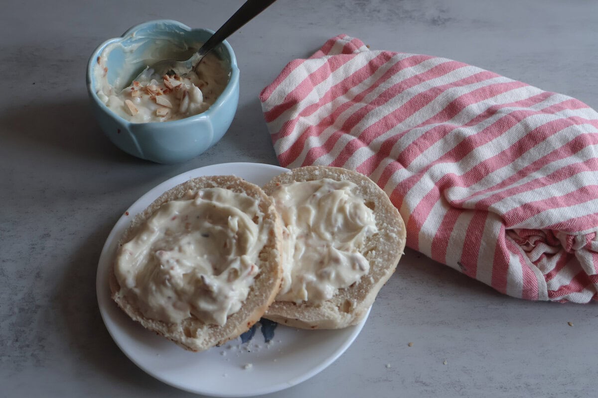 Honey almond cream cheese on a bagel, cream cheese in a blue bowl, pink and white striped tea towel.