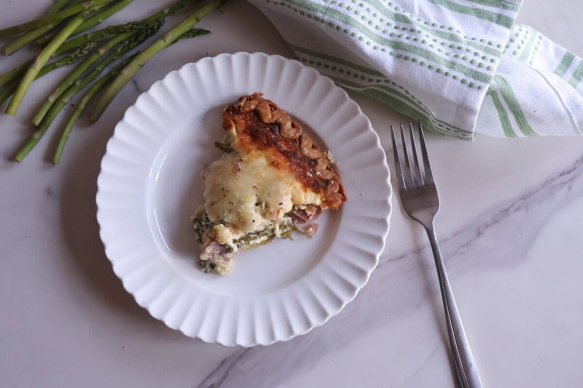 Slice of quiche on a white plate with a fork and a grenn and white tea towel.