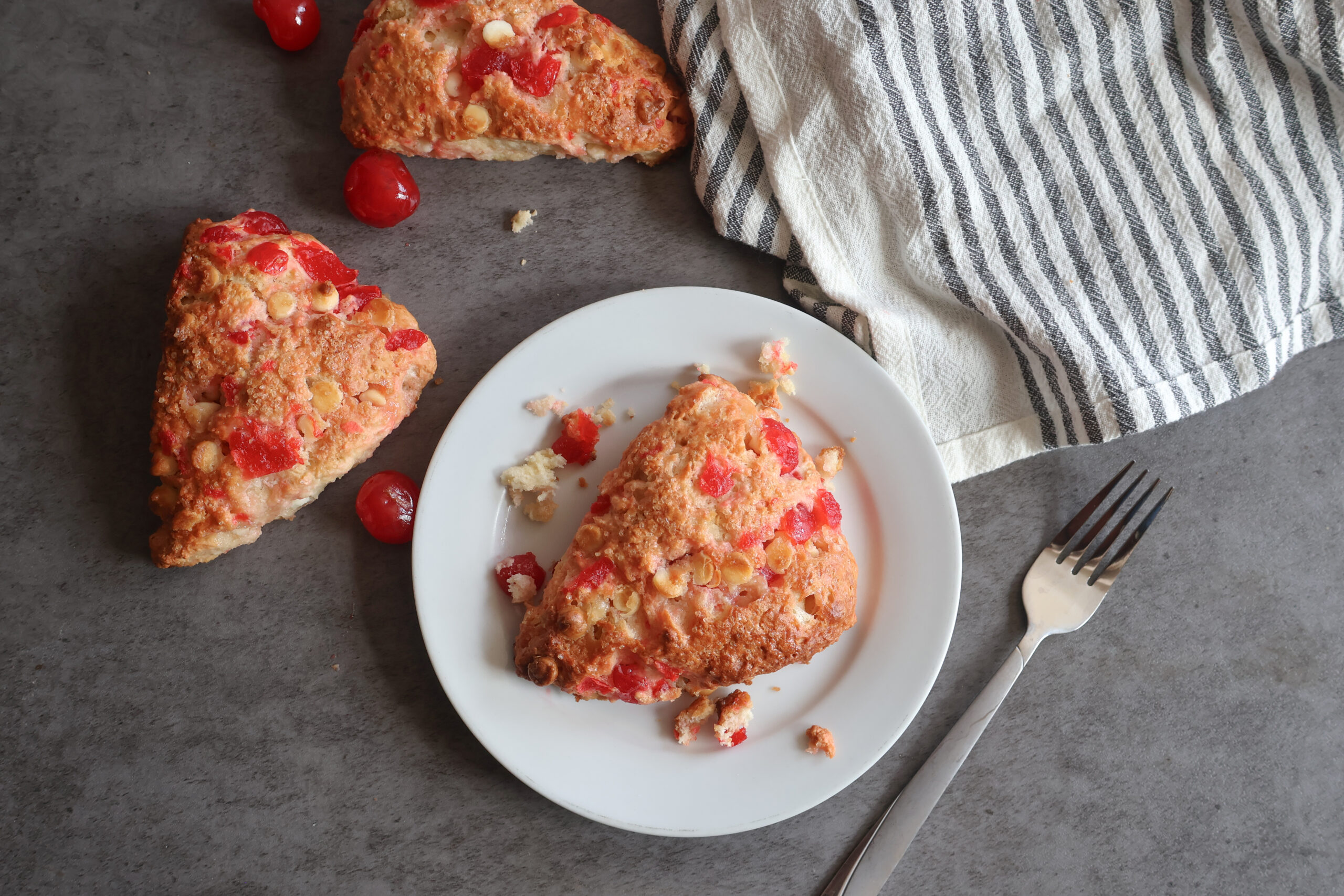 Cherry white chocolate scones on a white plate with a fork and a gray striped tea towel.