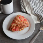 maraschino cherry white chocolate scone on a white plate with a fork, a gray striped tea towel and a cup of coffee.