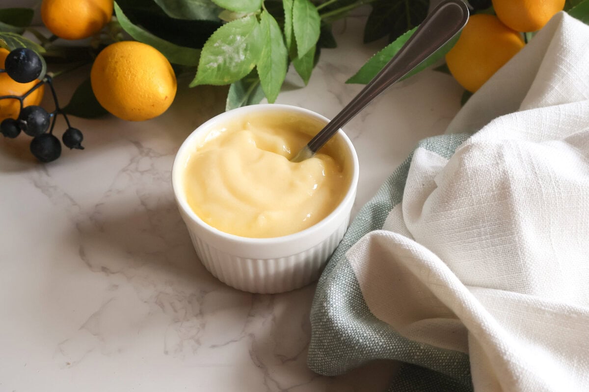 Lemon curd in a white dish with a spoon and a tea towel with lemon and leafs.