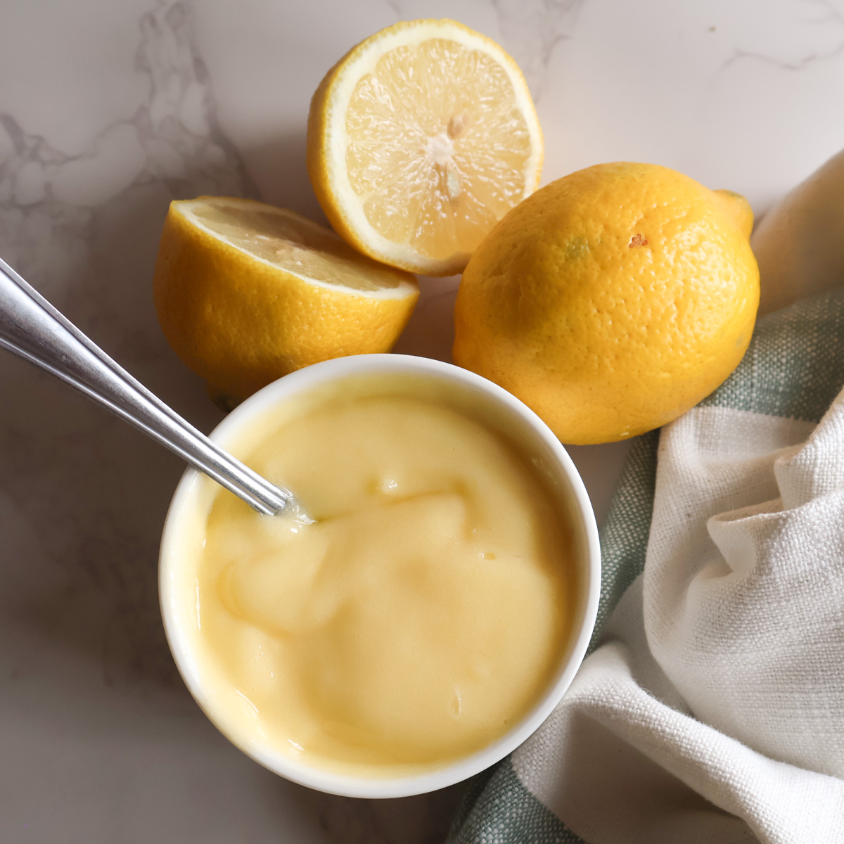Homemade lemon curd in a small white dish with a spoon and fresh lemons.