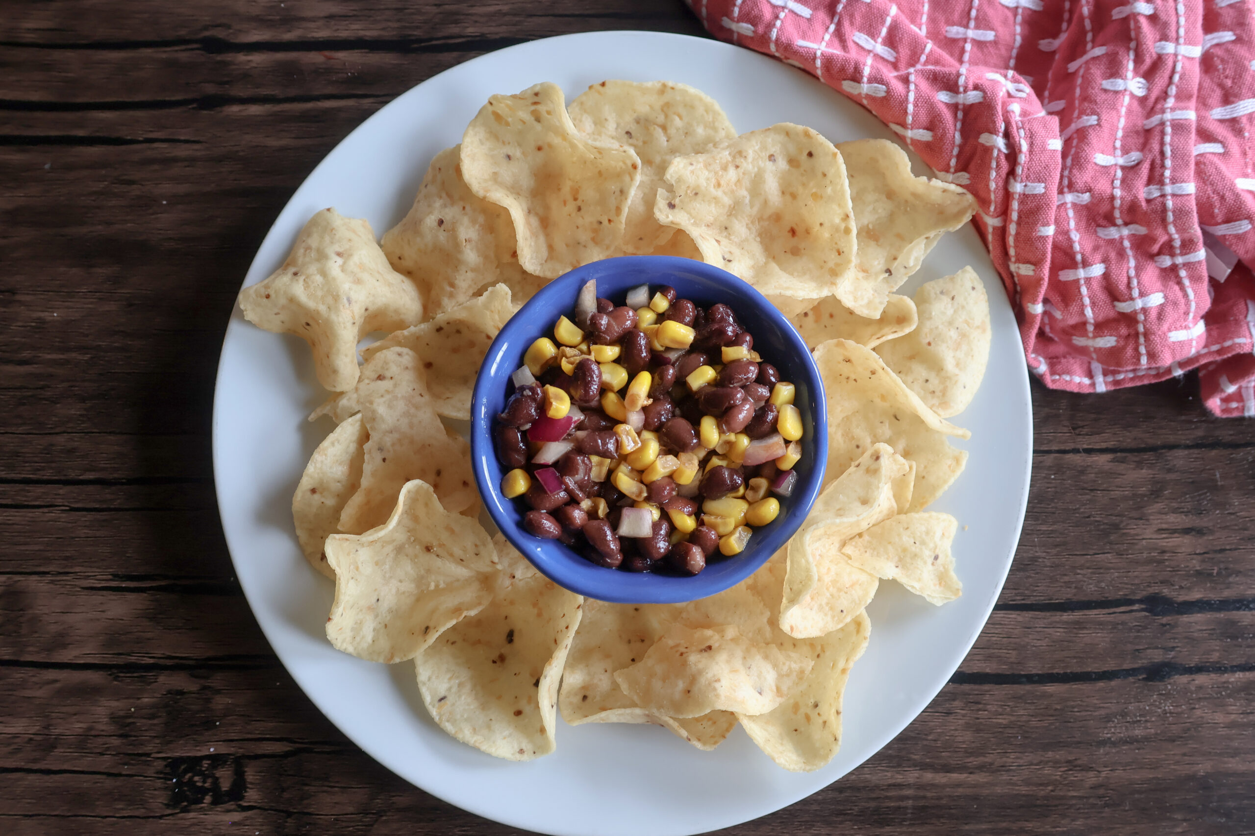 corn and black bean dip in a blue ramekin on a white plate with chips and a pink tea towel.