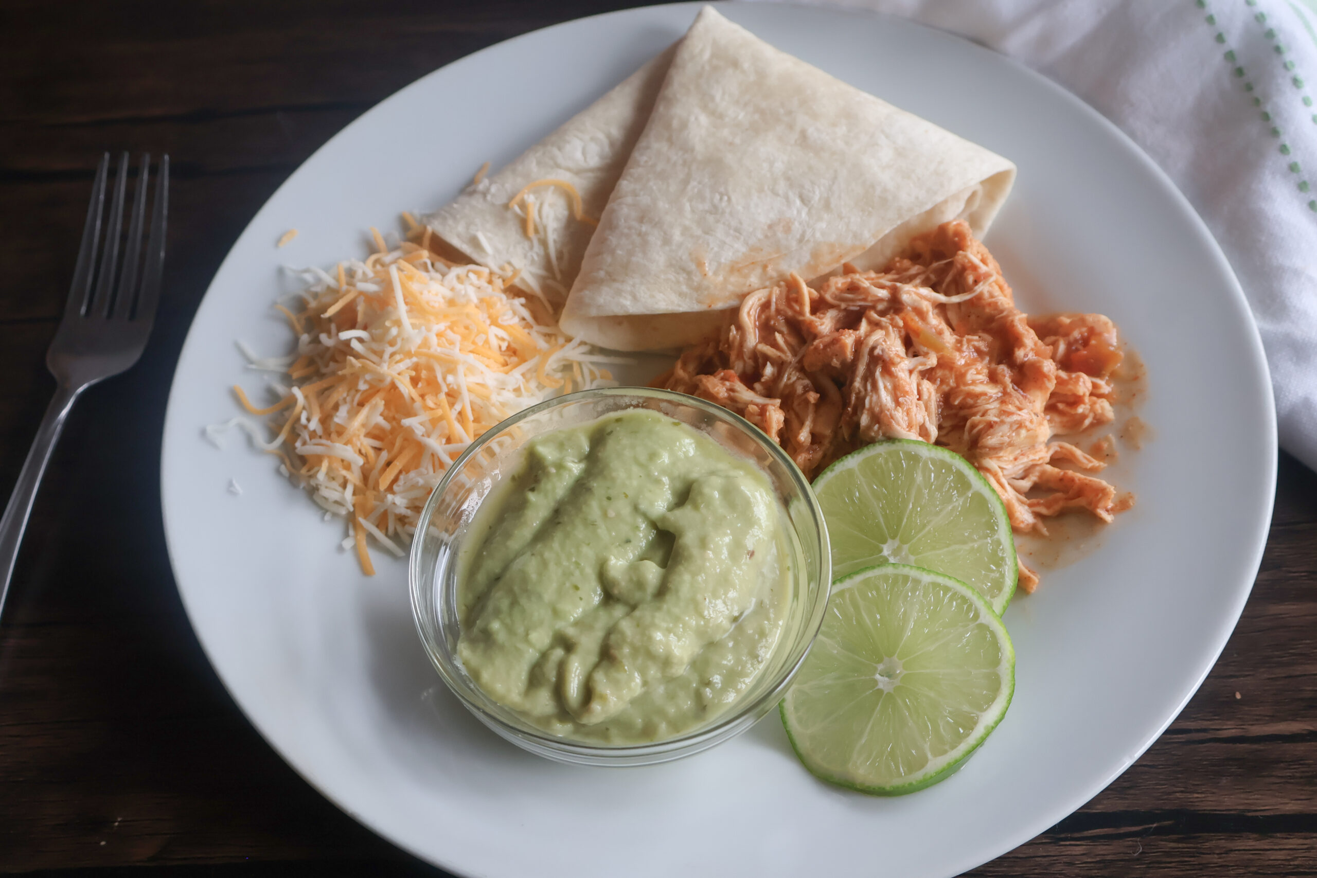 crockpot chicken tacos on a white plate with cheese, guacamole, and lime.