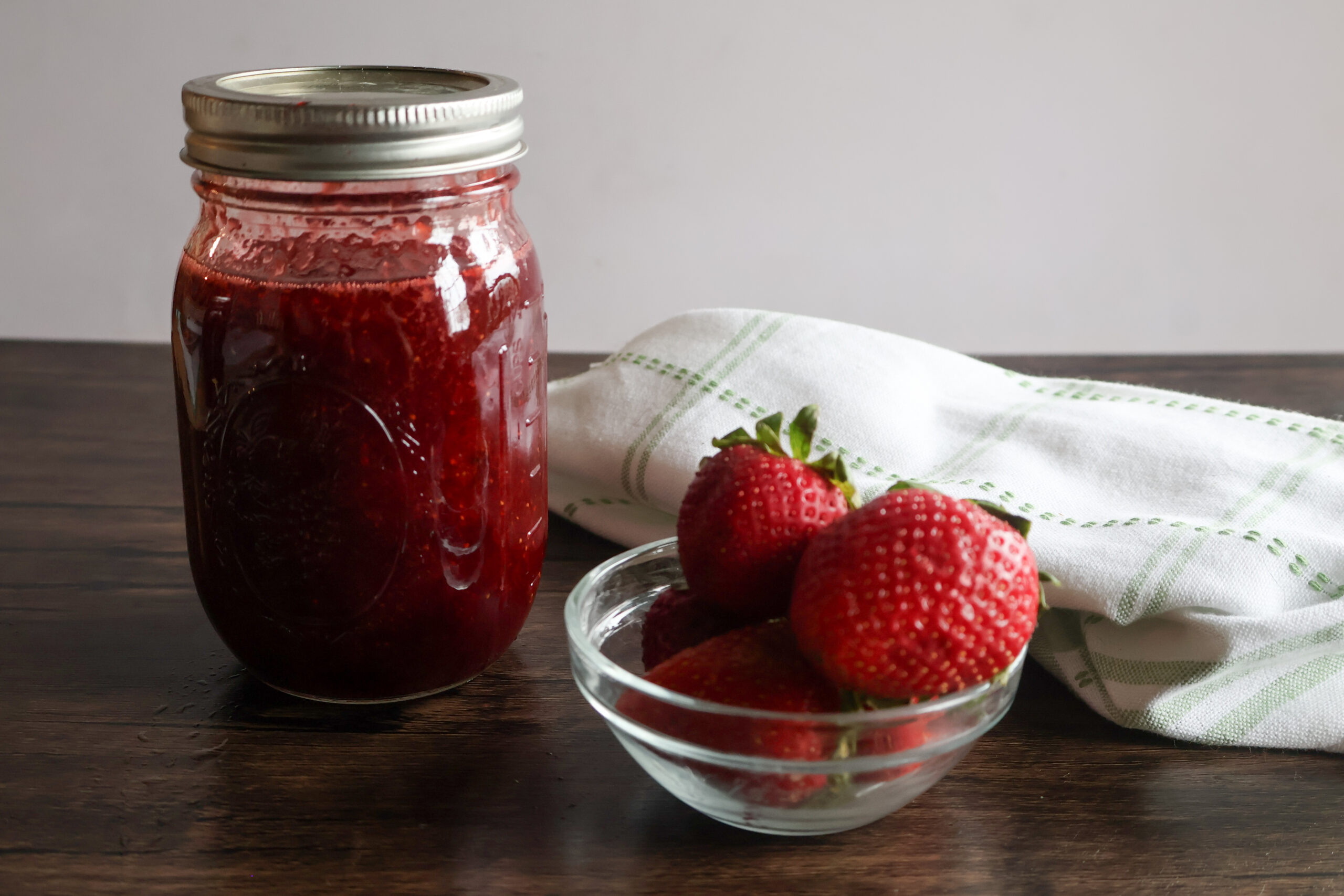 Homemade honey strawberry preserves in a jar with a small dish of strawberrues and a tea towel.
