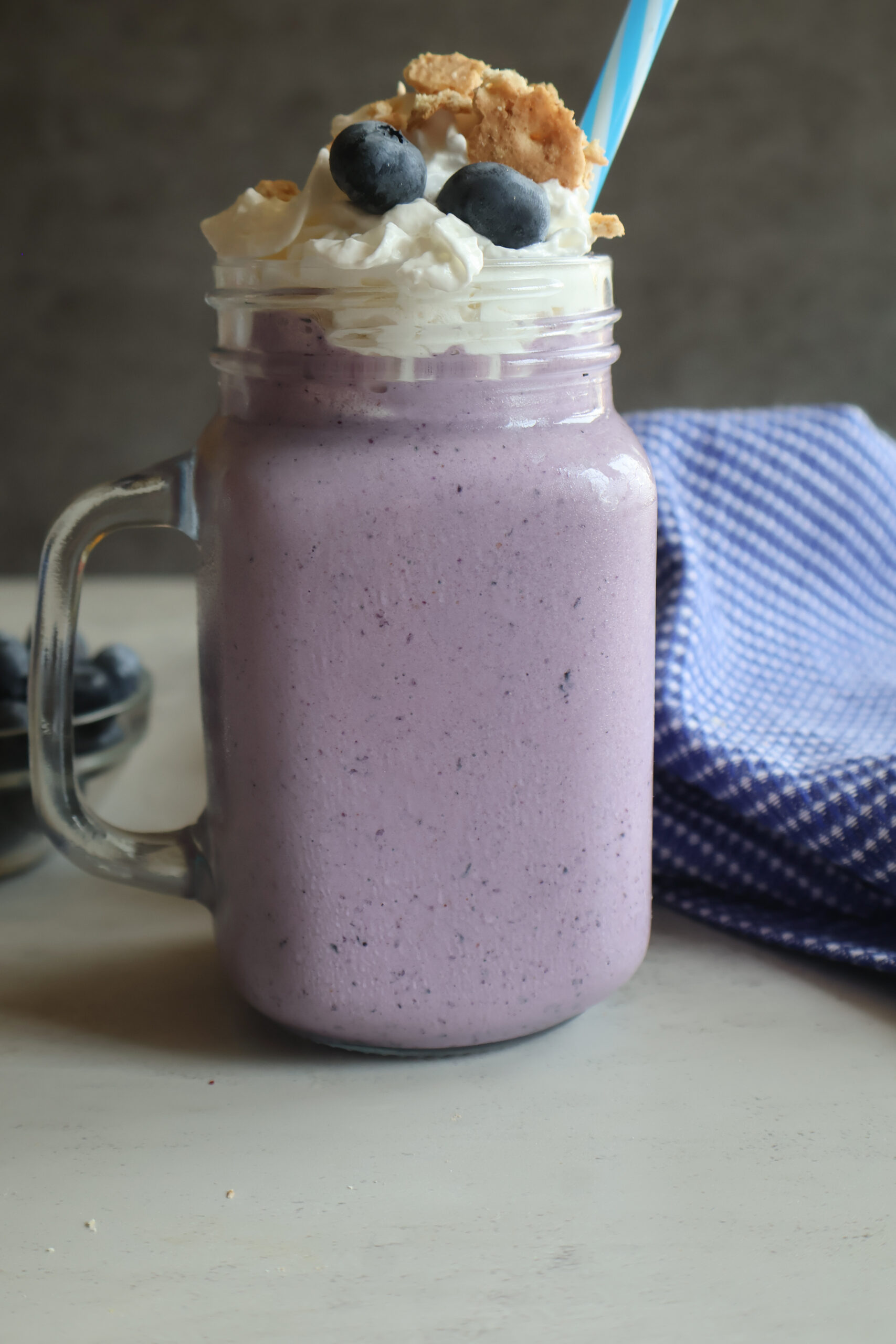 blueberry cheesecake milkshake in a glass topped with whipped cream, fresh blueberries and crushed graham crackers.