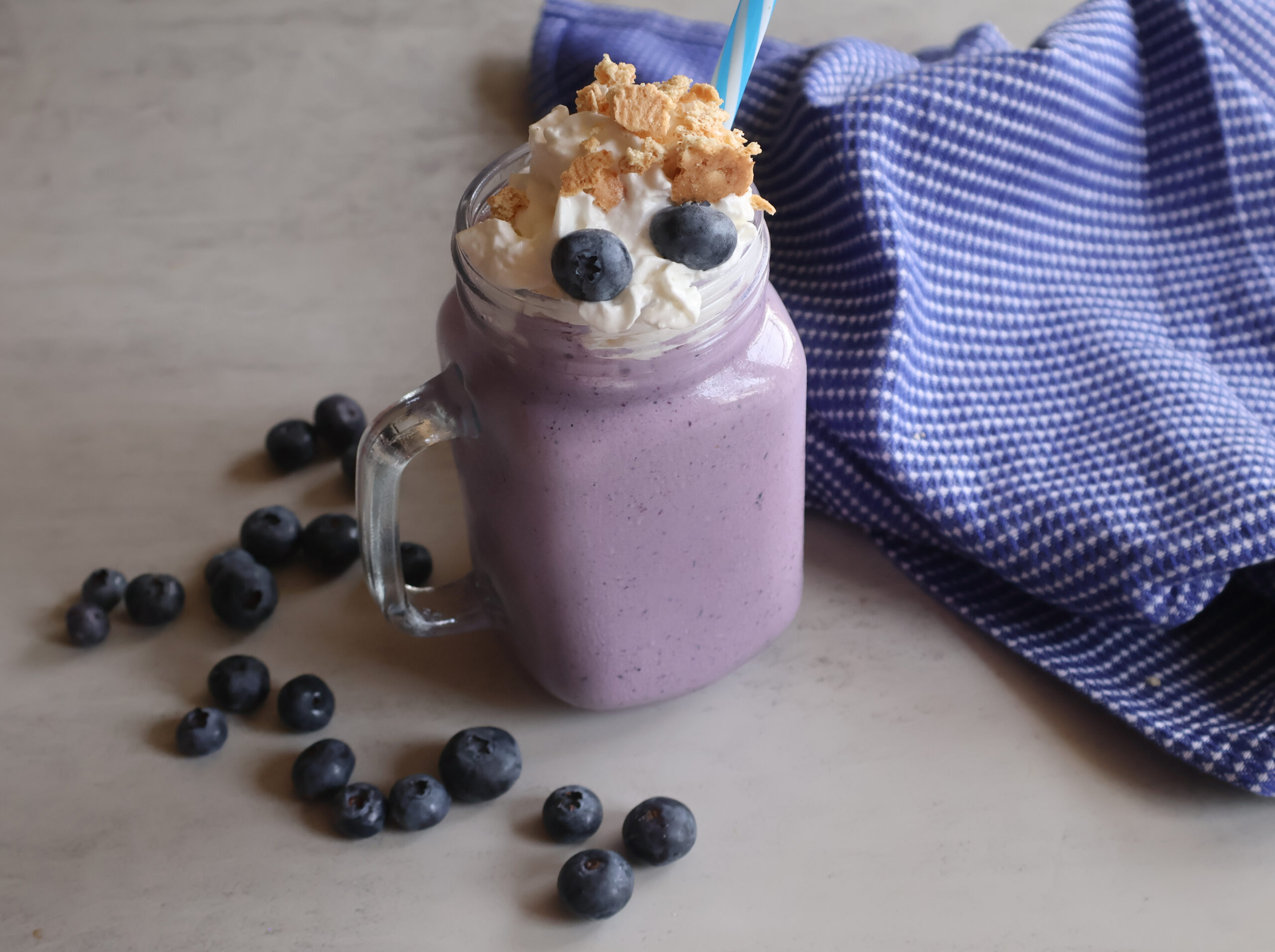 Blueberry Cheesecake Milkshake in a glass with a blue tea towel and fresh blueberries.