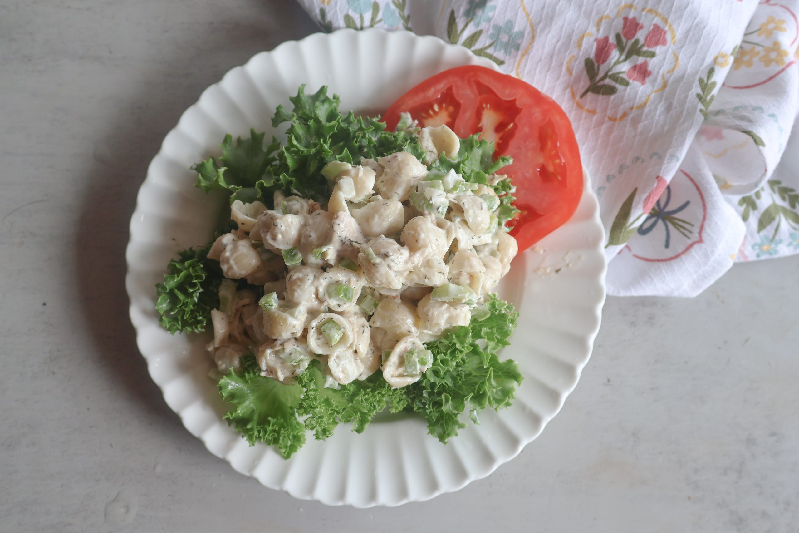 Creamy Tuna Pasta Salad on a white plate with lettuce and tomato with a tea towel.