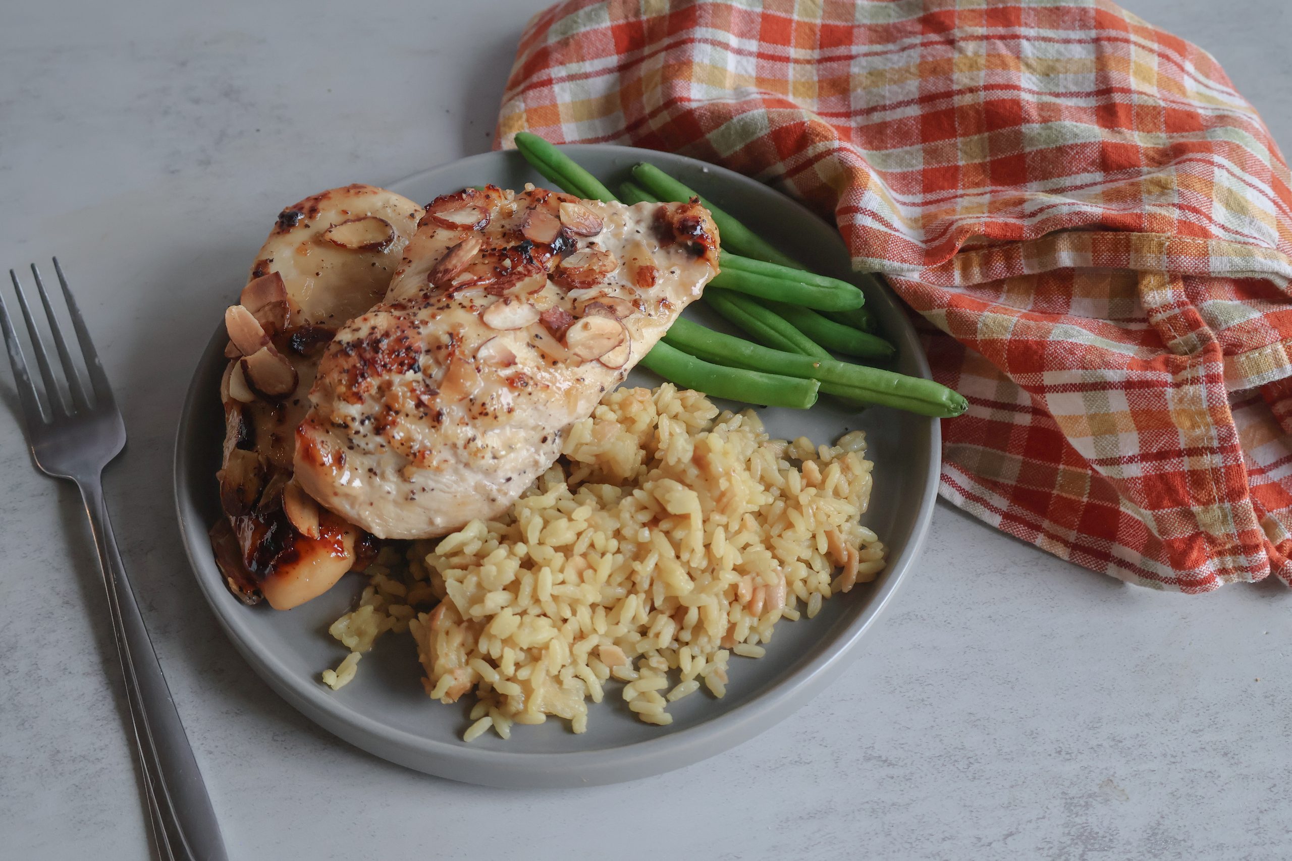 Vidalia onion almond chicken on a gray plate with green beans and rice with an orange plaid tea towel and a fork.