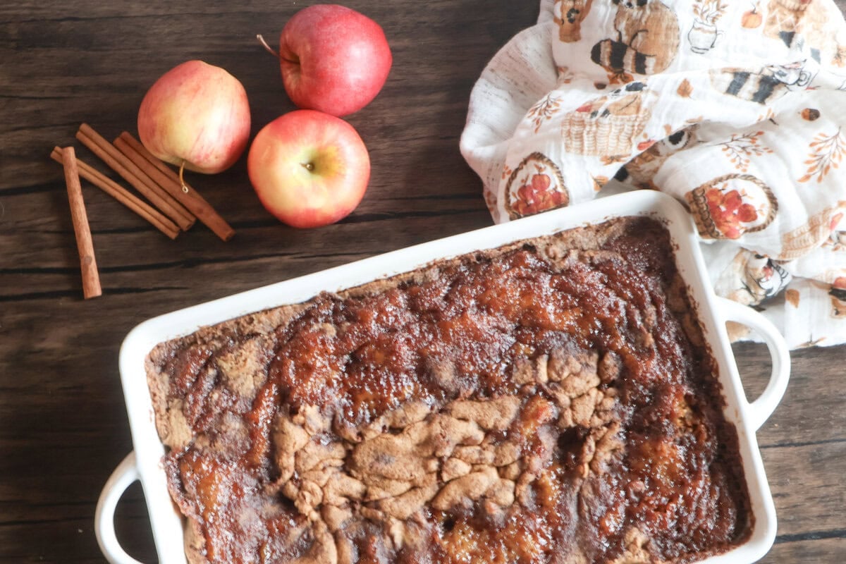 Apple cake in a white casserole dish with apples, cinnamon sticks, and an apple tea towel.