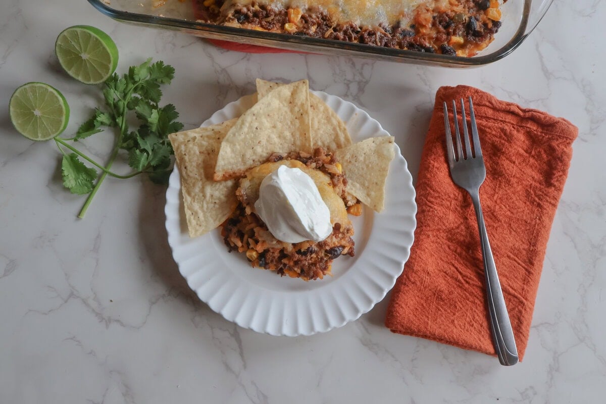 Serving of cheesy mexican beef and rice casserole on a white plate with tortilla chips. lime, cilantro and a fork.